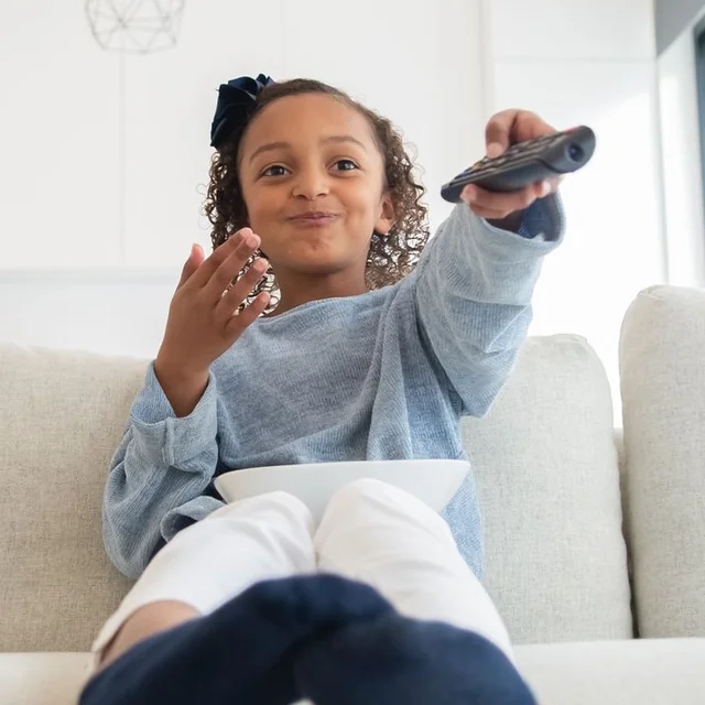 Une petite fille assise sur un canapé pointe la télécommande vers la télévision